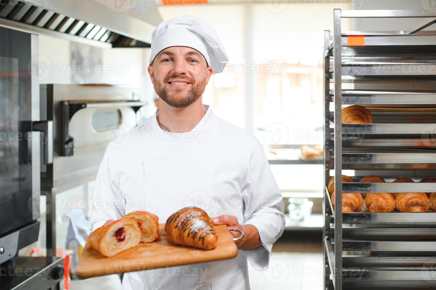 Smiling baker holding tray with fresh cooked croissants in bakehouse photo