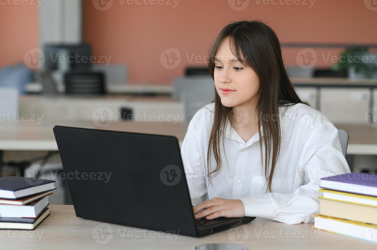 A beautiful caucasian female student is studying in college remotely. She is sitting with a laptop and a notepad and concentrated is watching a conference lesson photo