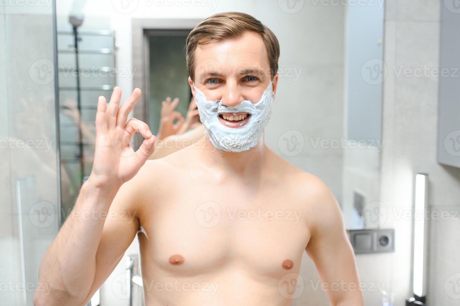 Handsome young man is shaving his face and looking at the mirror. photo