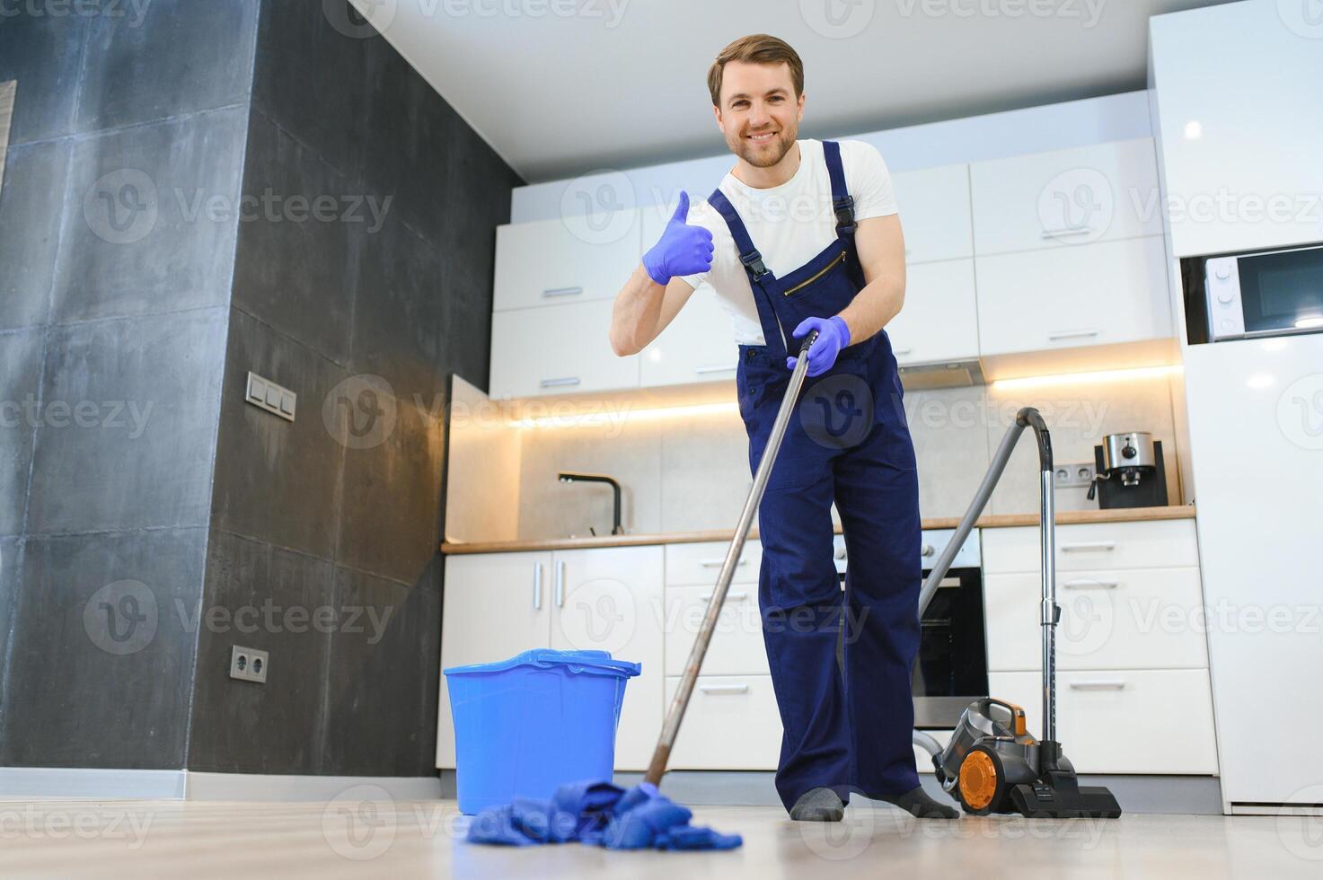 Professional cleaner in blue uniform washing floor and wiping dust from the furniture in the living room of the apartment. Cleaning service concept photo