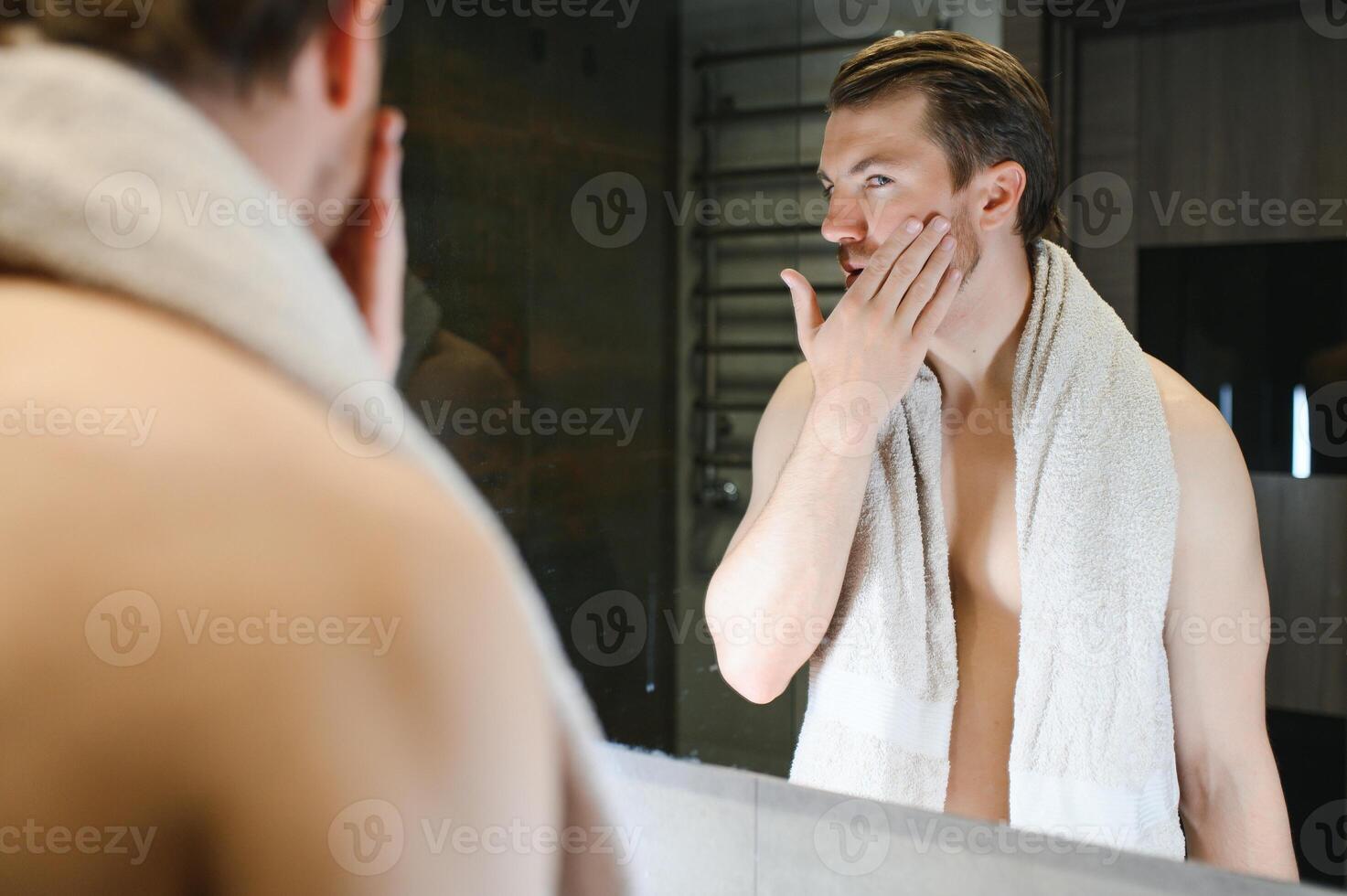 Young Caucasian man look in mirror do skincare facial routine after shower in home bathroom, millennial male in towel after bath apply face lotion or cream on skin after shaving, hygiene concept. photo