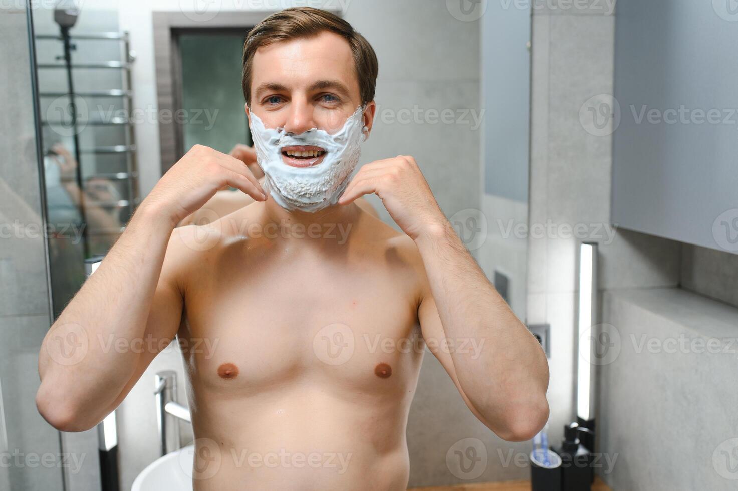 Young smiling Caucasian shirtless man applying shaving foam on face in front of mirror, preparing for hair removal morning routine. Concept of personal hygiene, skincare, male facial beauty products. photo