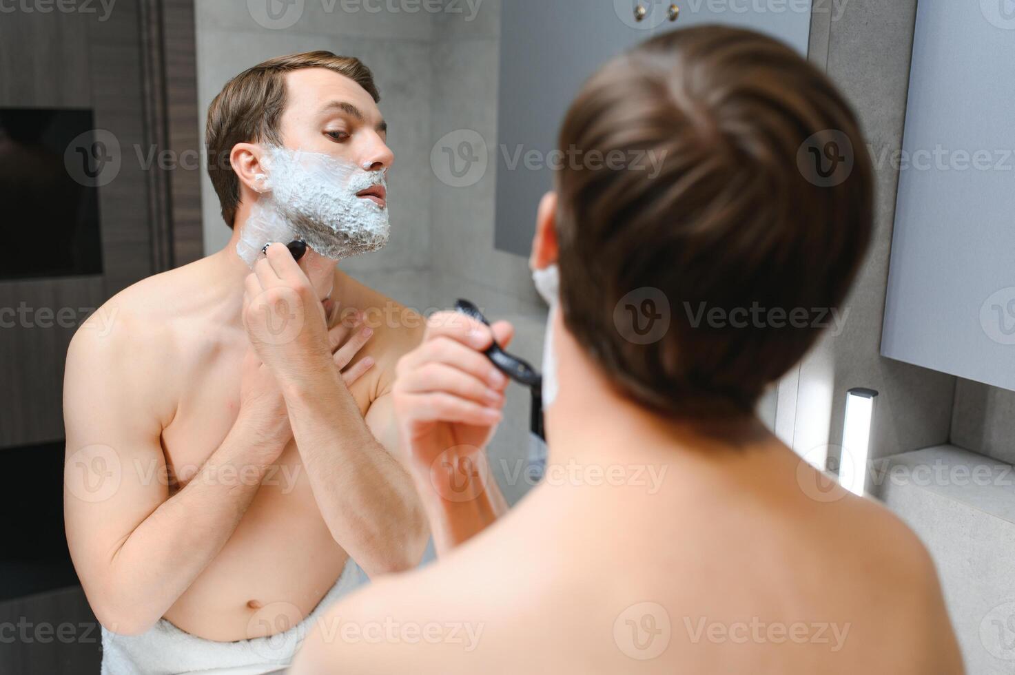 Handsome young man is shaving his face and looking at the mirror. photo