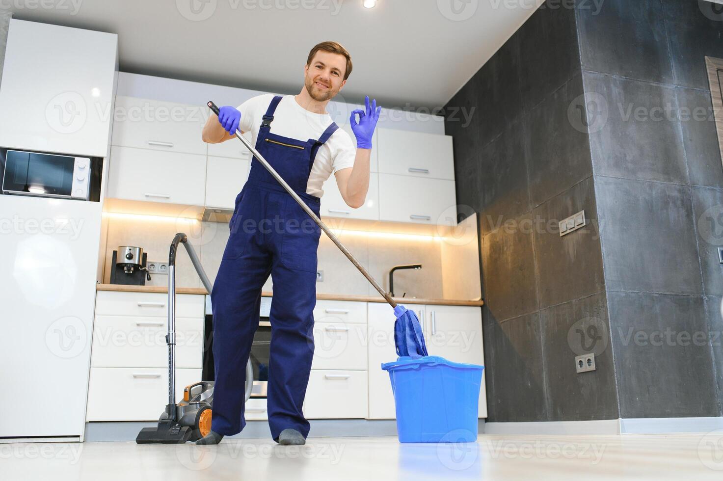 Professional cleaner in blue uniform washing floor and wiping dust from the furniture in the living room of the apartment. Cleaning service concept photo