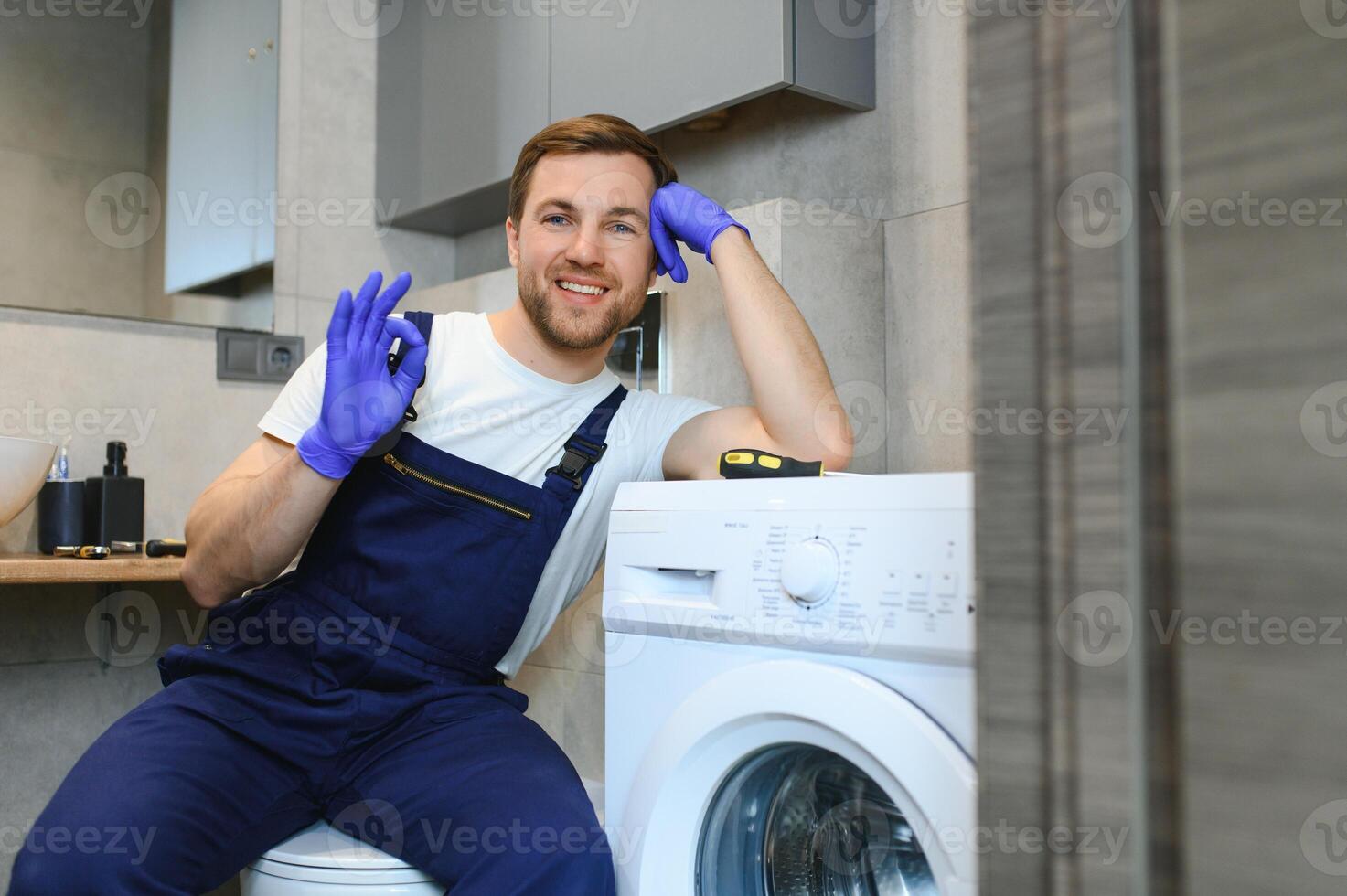 The young handsome repairman in worker suit with the professional tools box is fixing the washing machine in the bathroom photo