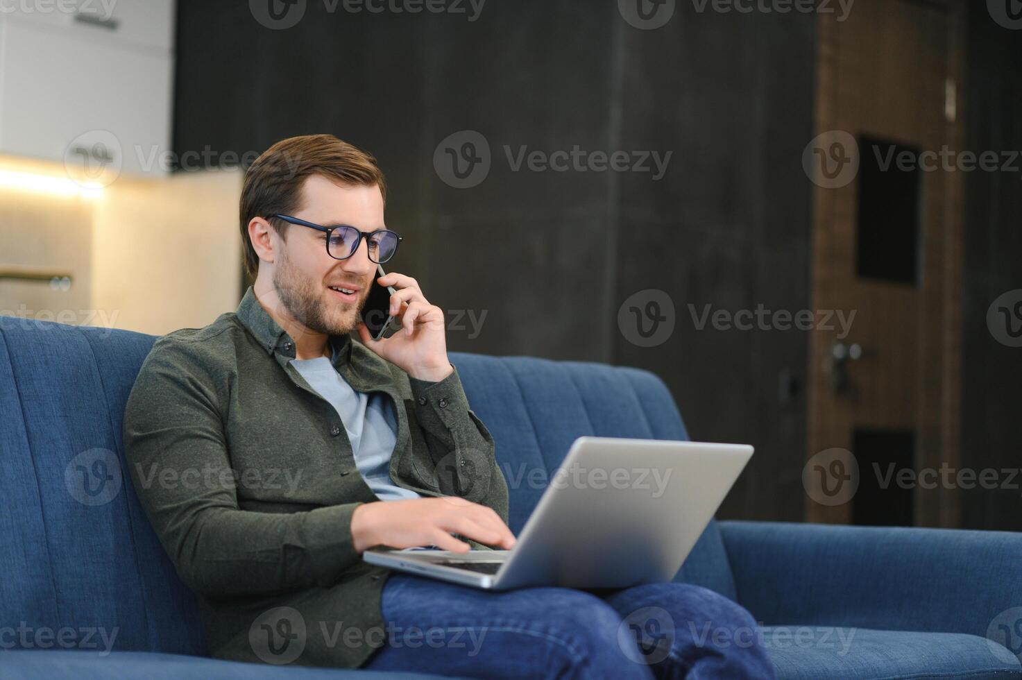 young freelancer man in stylish glasses working at laptop computer, sitting on floor at home, using wireless technology for job, leisure on Internet, thinking, touching chin photo