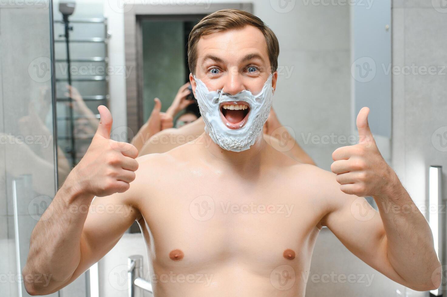 Young smiling Caucasian shirtless man applying shaving foam on face in front of mirror, preparing for hair removal morning routine. Concept of personal hygiene, skincare, male facial beauty products. photo