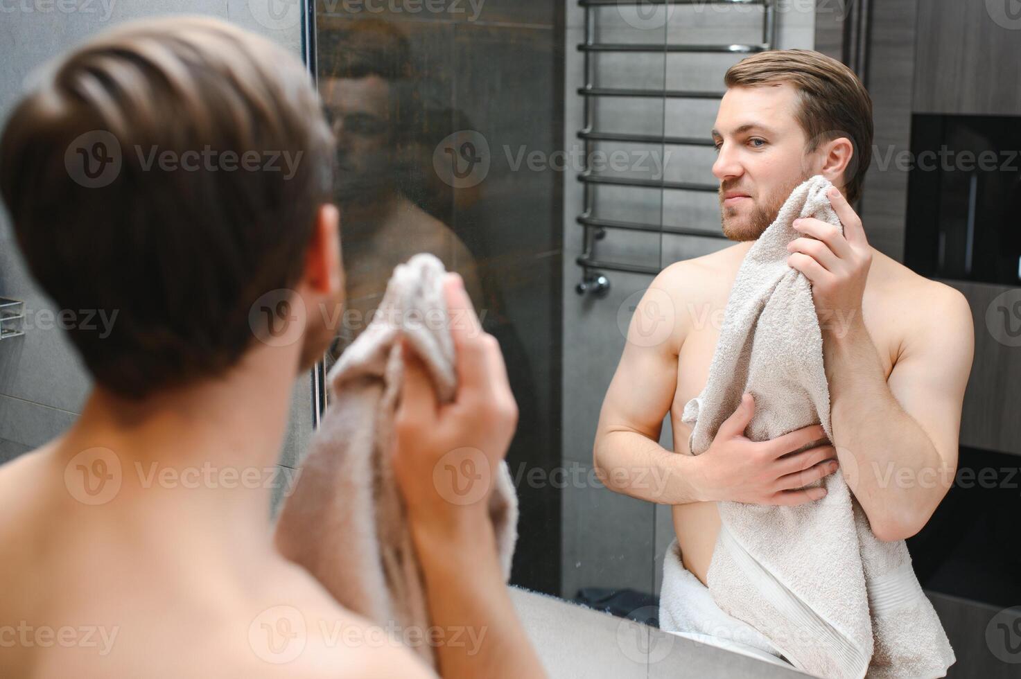 Young Caucasian man look in mirror do skincare facial routine after shower in home bathroom, millennial male in towel after bath apply face lotion or cream on skin after shaving, hygiene concept. photo