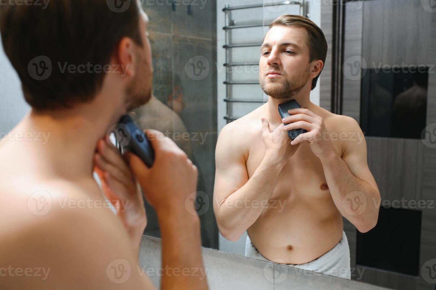 beauty, shaving, grooming and people concept - close up of young man looking to mirror and shaving beard with trimmer or electric shaver at home bathroom photo