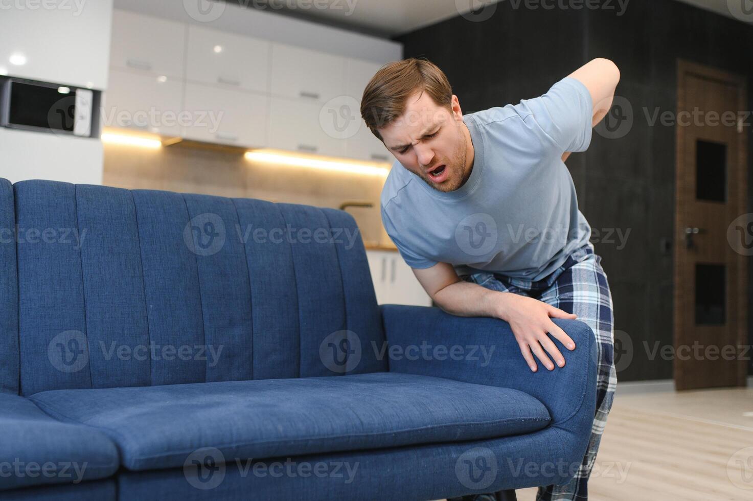 People, Healthcare and Treatment. Portrait of sick man suffering from acute side back pain, sitting on couch at home, person touching lower back aching area with hand. Health Problem Concept photo
