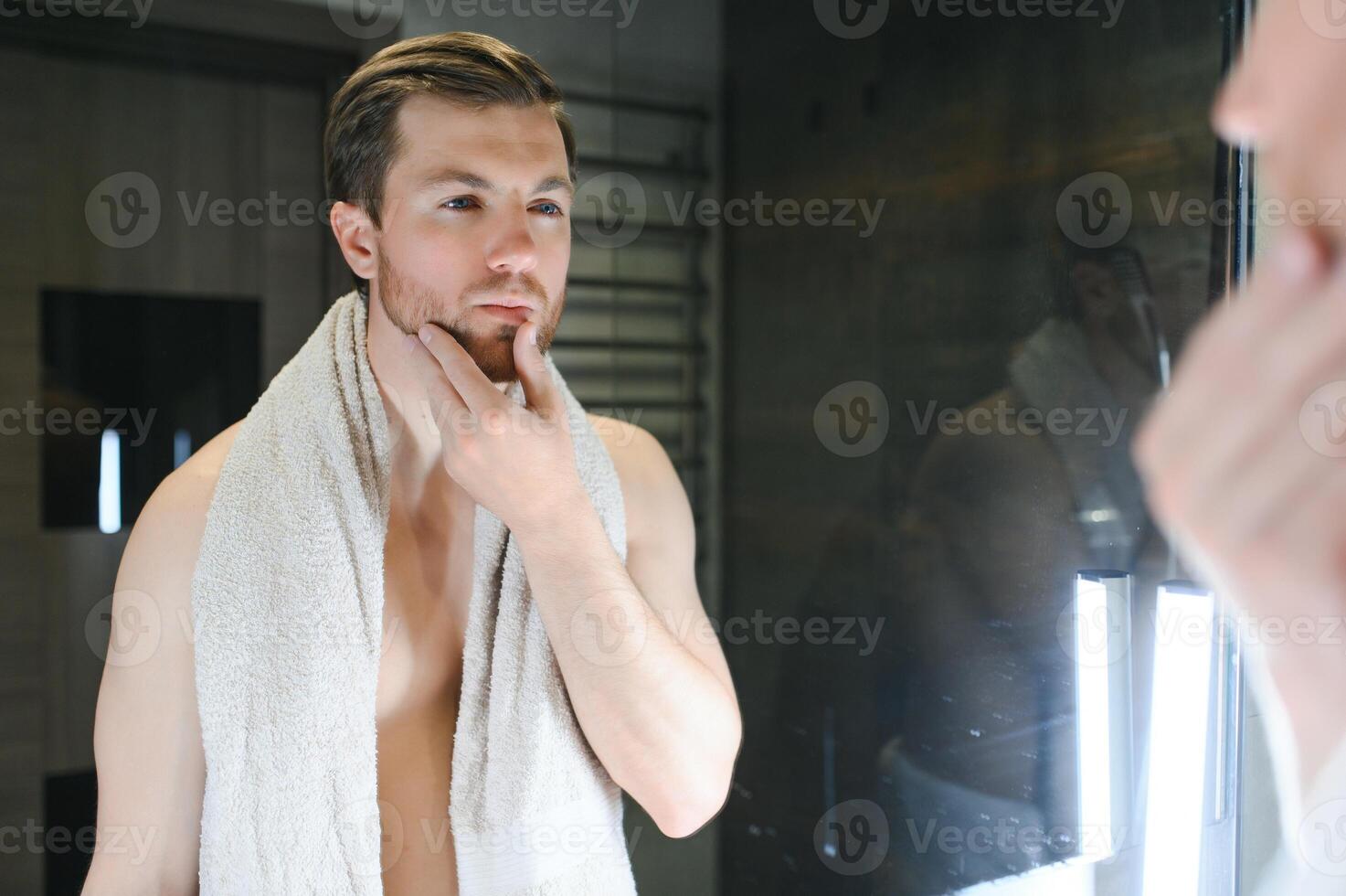 Young Caucasian man look in mirror do skincare facial routine after shower in home bathroom, millennial male in towel after bath apply face lotion or cream on skin after shaving, hygiene concept. photo