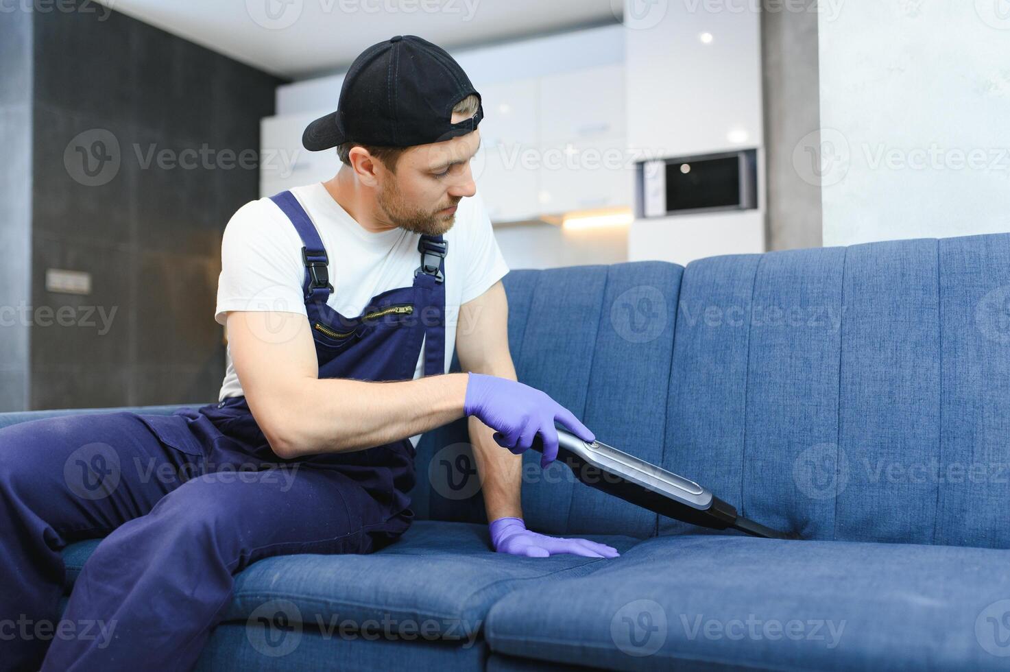 Cleaning service. Man janitor in gloves and uniform vacuum clean sofa with professional equipment. photo
