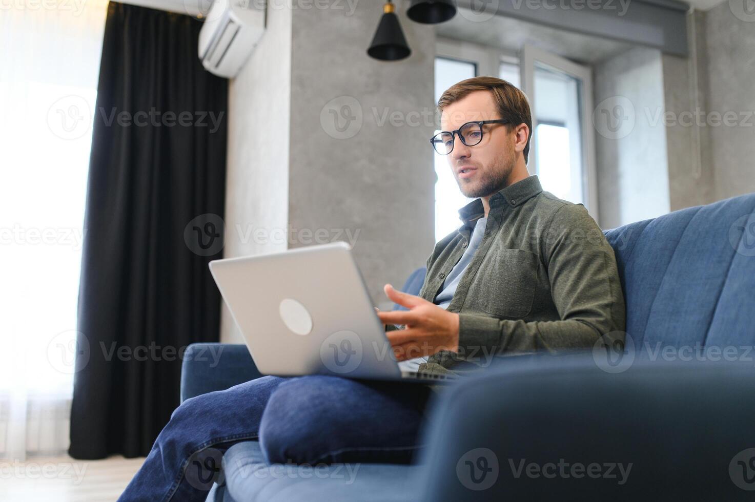 young freelancer man in stylish glasses working at laptop computer, sitting on floor at home, using wireless technology for job, leisure on Internet, thinking, touching chin photo
