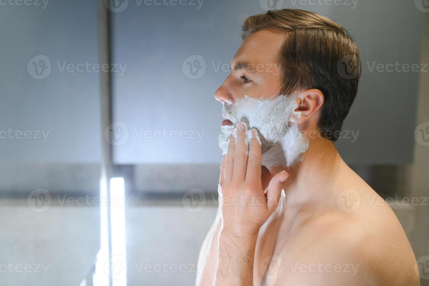 Half is done. Handsome youthful topless man is applying mean of shaving on his face. photo