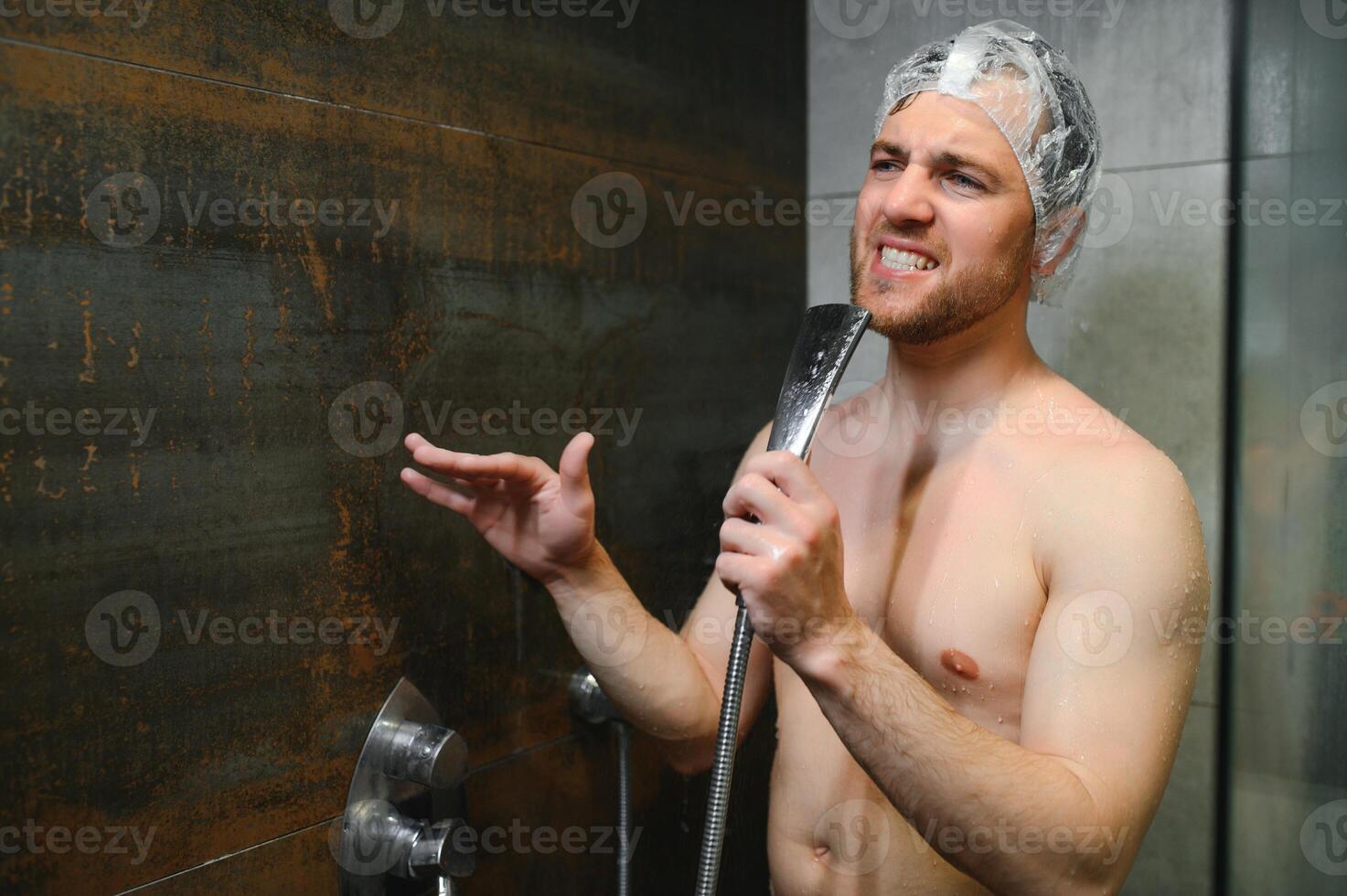 Attractive young cheerful man singing while washing in the shower, wearing shower cap and holding shower head photo