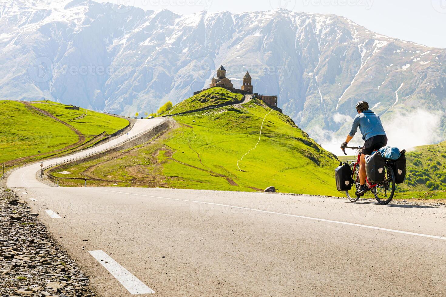 Male cyclist on touring bicycle cycling towards Gergeti trinity church with mountains background. Cycling holidays and travel around caucasus mountain range. photo