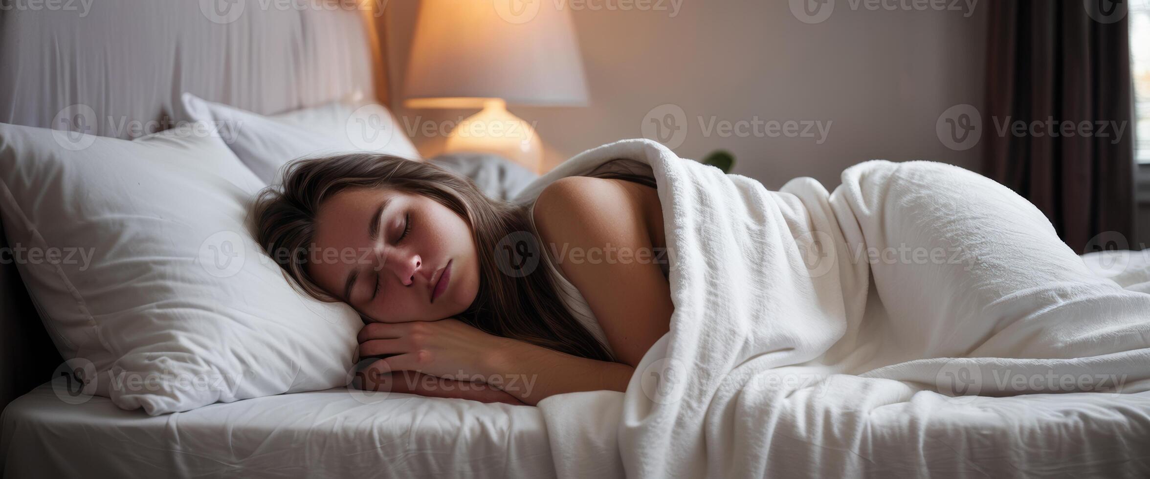 A young woman with long brown hair sleeps peacefully in a cozy bed, wrapped in white bedding. The serene bedroom setting exudes comfort and tranquility. photo