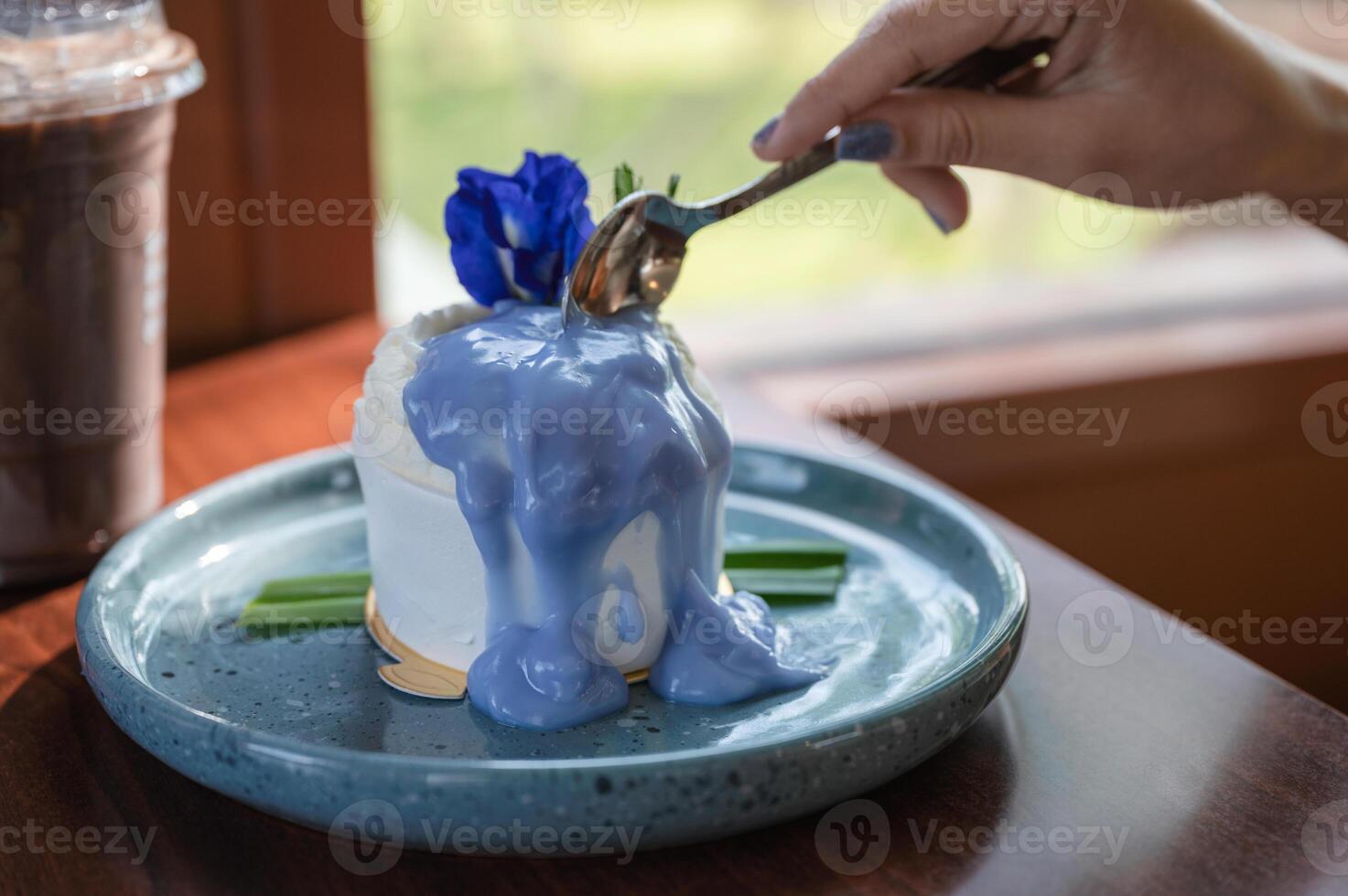 Woman hand using spoon cutting coconut cake with butterfly pea topping on ceramic dish and iced cocoa on the table photo