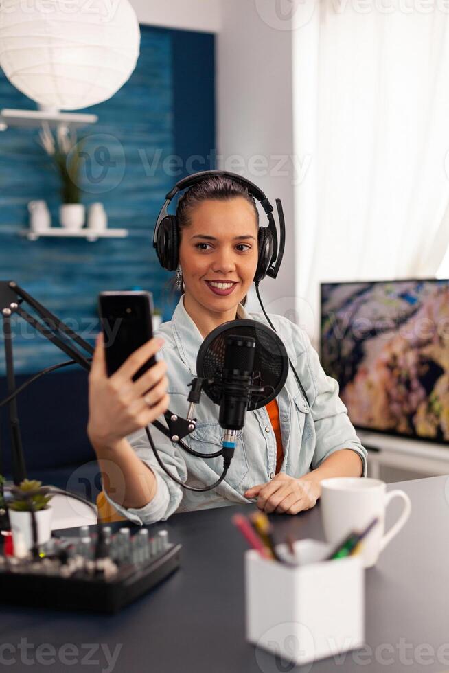 Portrait of blogger woman taking selfie for audience using smartphone working in podcast home studio. Content creator recording new fashion and beauty review and having fun on social media platform. photo