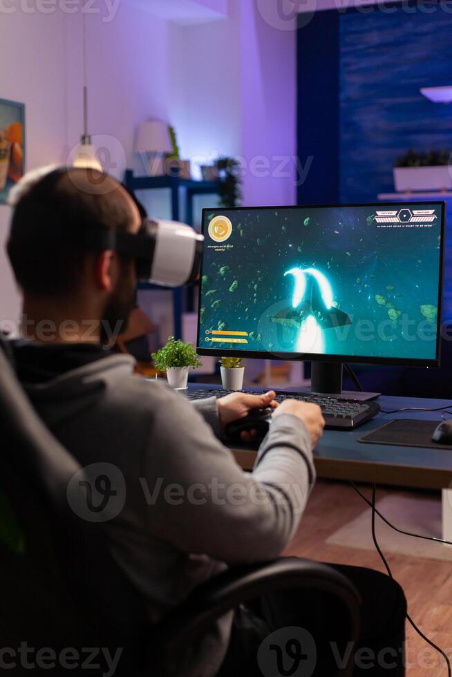 Player man playing game at powerful computer late night wearing vr headset. Excited player using wireless controller for virtual tournament gaming space shooter at home photo