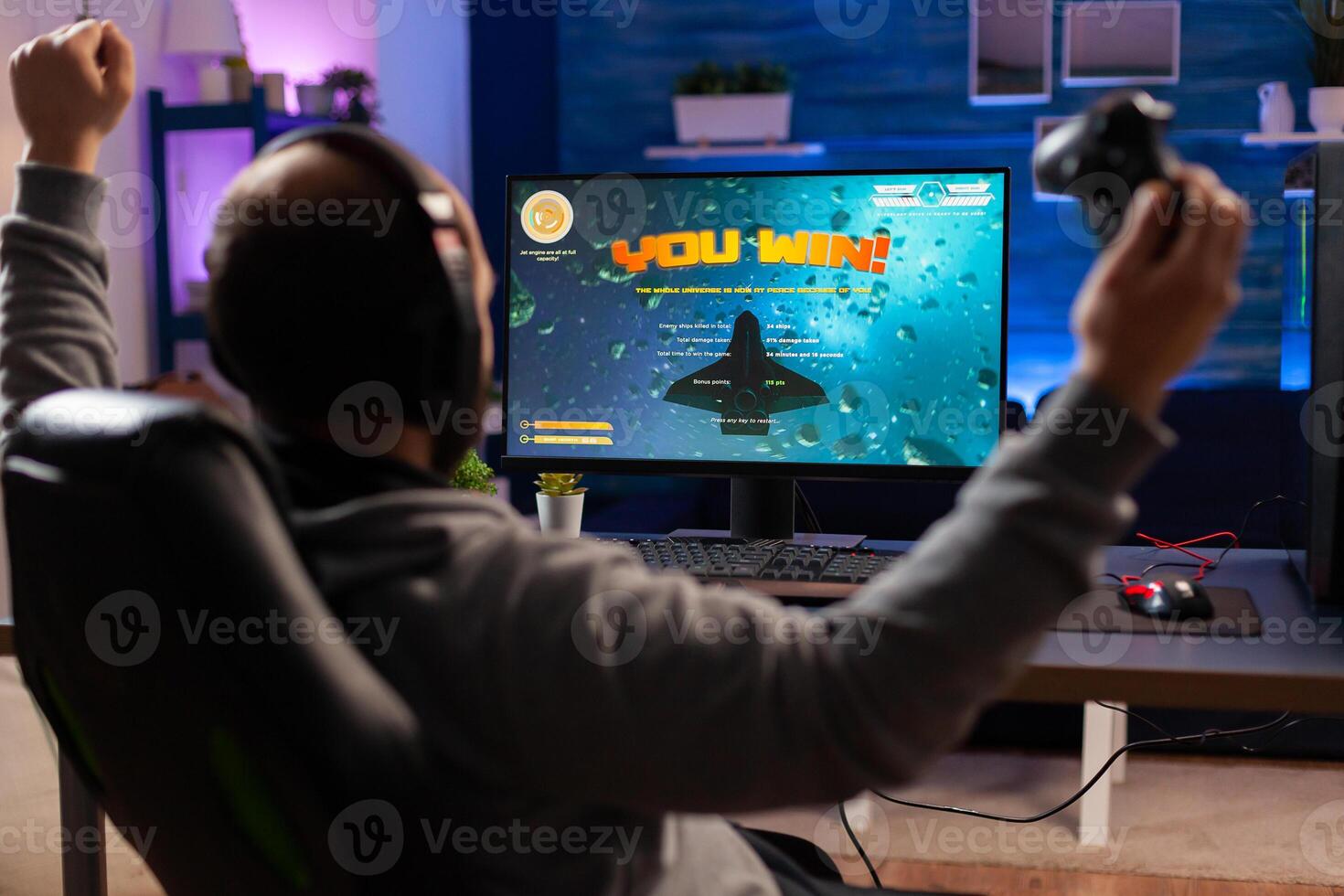 Addicted man playing space shooter videogame using headphones and wireless console. Excited gamer raising hands after winning online championship game at home gaming studio photo