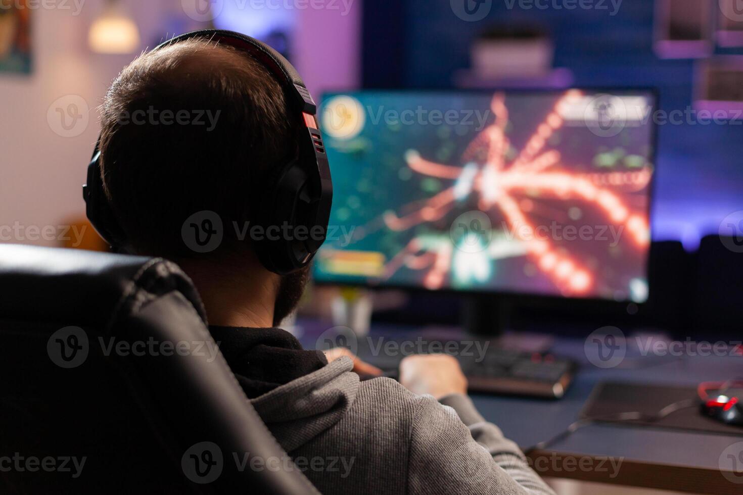 Back view shot of concentrate gamer streaming online videogames on computer using wireless controller. Player man with headphones playing games in room with neon light and professional equipment photo