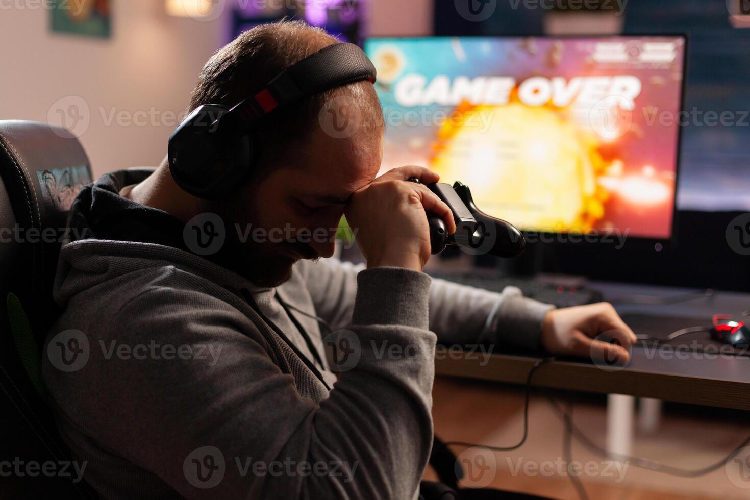 Upset cyber player losing game sitting on gaming desk late at night in living room. Man streaming games for online championship using headphones and professional joystick photo