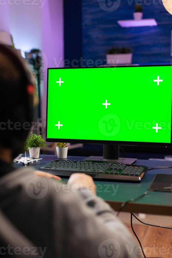 Gamer man playing videogames on computer with green screen holding wireless joystick in professional gaming studio. Player using pc with mock up isolated desktop streaming shooter games wearing headset photo