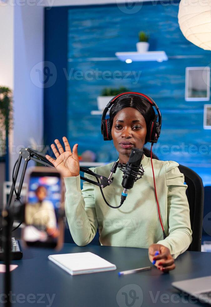 African blogger looking at camera and waving to camera recording episode for online show. Speaking during livestreaming, blogger discussing in podcast wearing headphones. photo