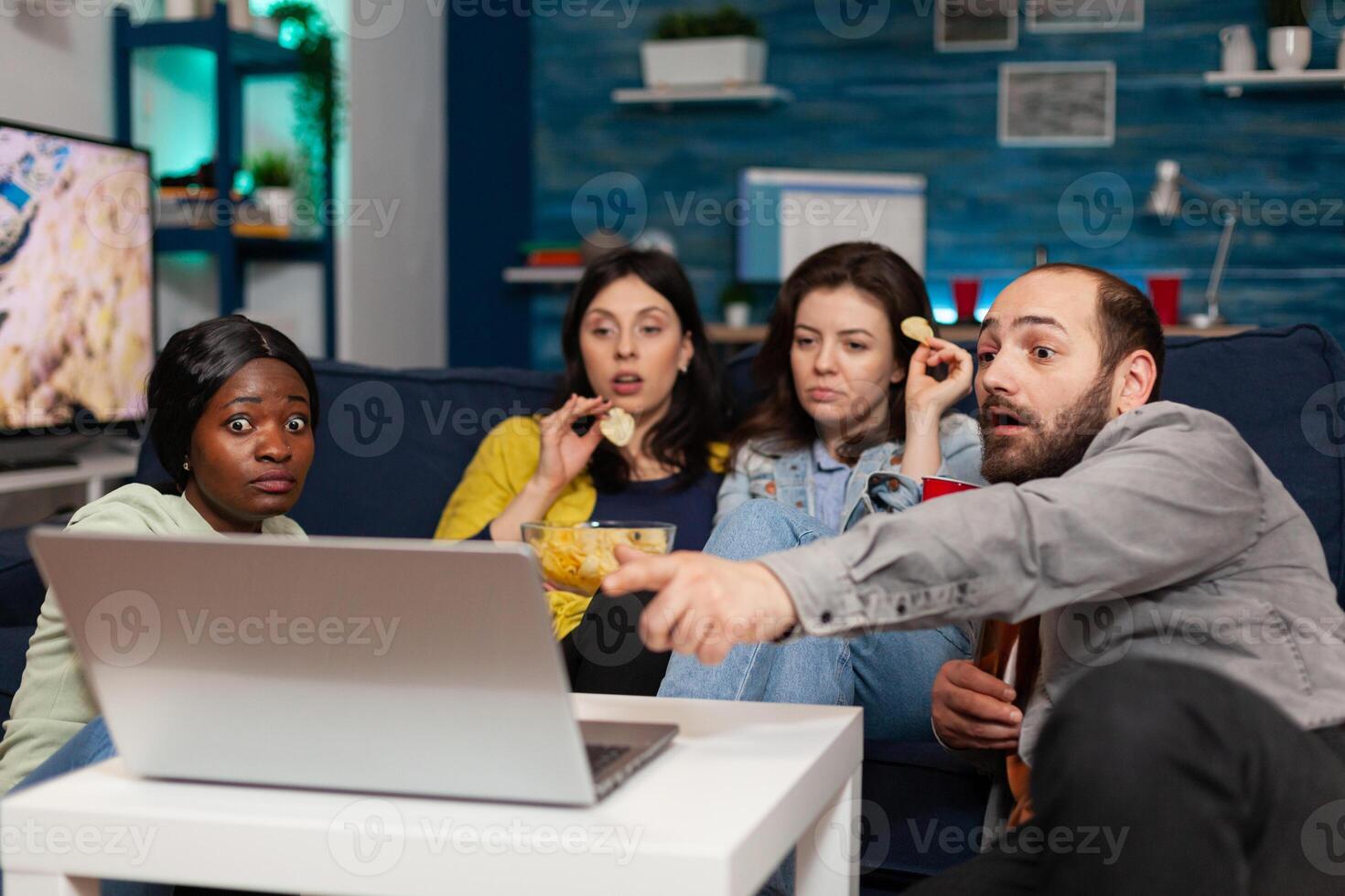 Multi-ethnic friends spending time together looking at entertainment on laptop talking about lifestyle. Group of multiracial people relaxing having fun chilling on movie night, hanging out photo