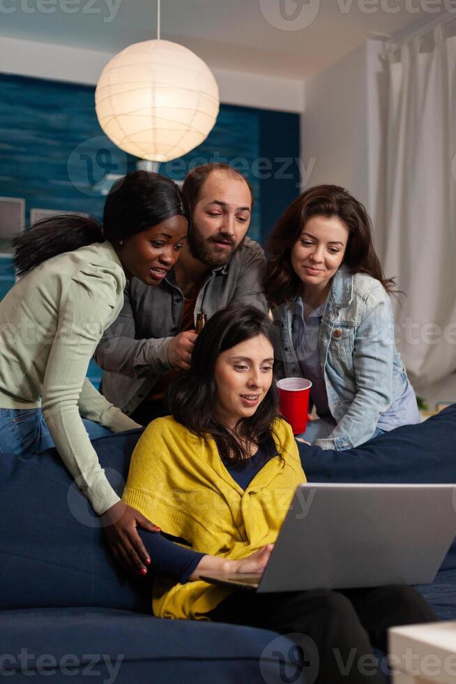 Multi ethnic friends hanging out talking on webcam laptop during call. Group of multiracial people spending time together sitting on couch late at night in living room. photo