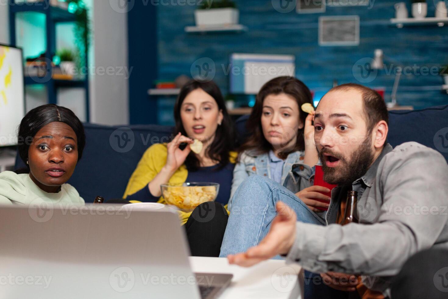 Multiracial friends relaxing on couch while watching comedy looking shocked during movie night. Group of multi-ethnic people spending time together drinking beer, eating snacks late at night. photo
