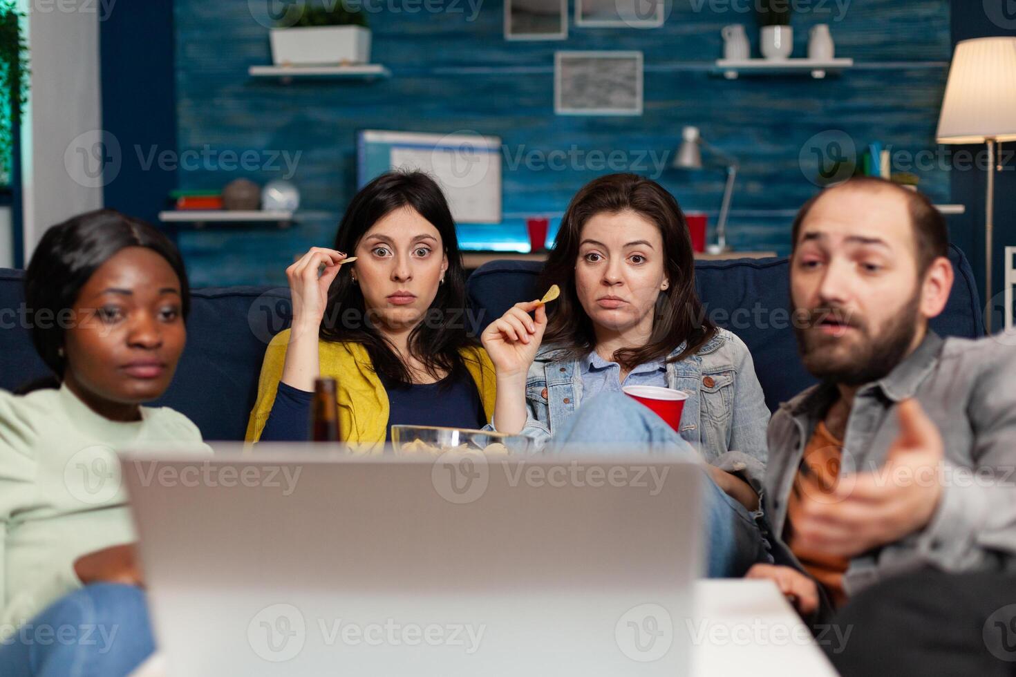 Multi-ethnic friends watching online film on laptop computer relaxing on couch together. Group of multiracial people hanging out, drinking beer, eating snacks late at night in living room. photo