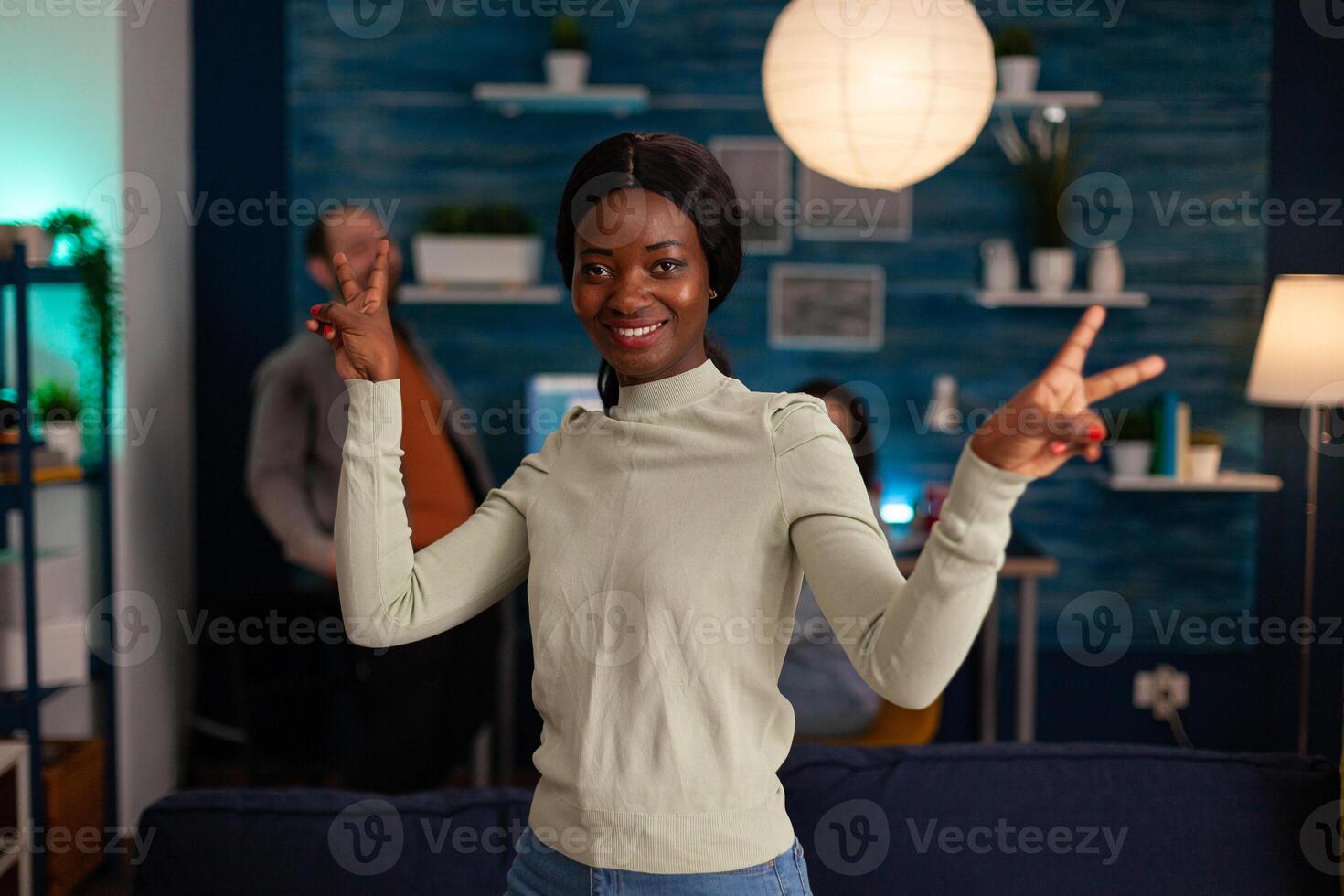 Potrait of afro american woman smiling into camera showing victory hand sign late at night in living room. In background multiethnic friends gathering together having fun during wekeend party. photo
