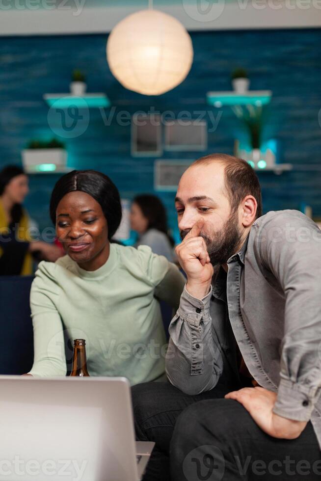 Multi ethnic friends socializing celebrating friendship watching funny on laptop sitting on couch. In background two women drinking beer enjoying time spend together during entertainment party. photo