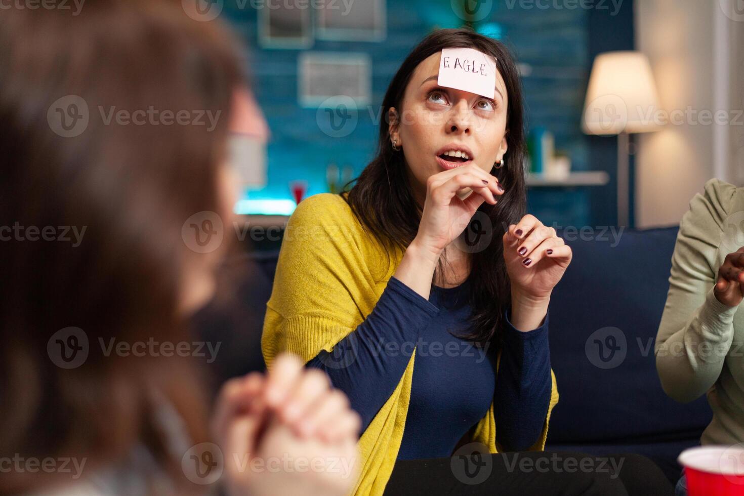 Mixed race friends with sticky notes attached on forehead playing guess who game during home party. Multi-ethnic people having fun, laughing together while sitting on sofa in living room late at night. photo