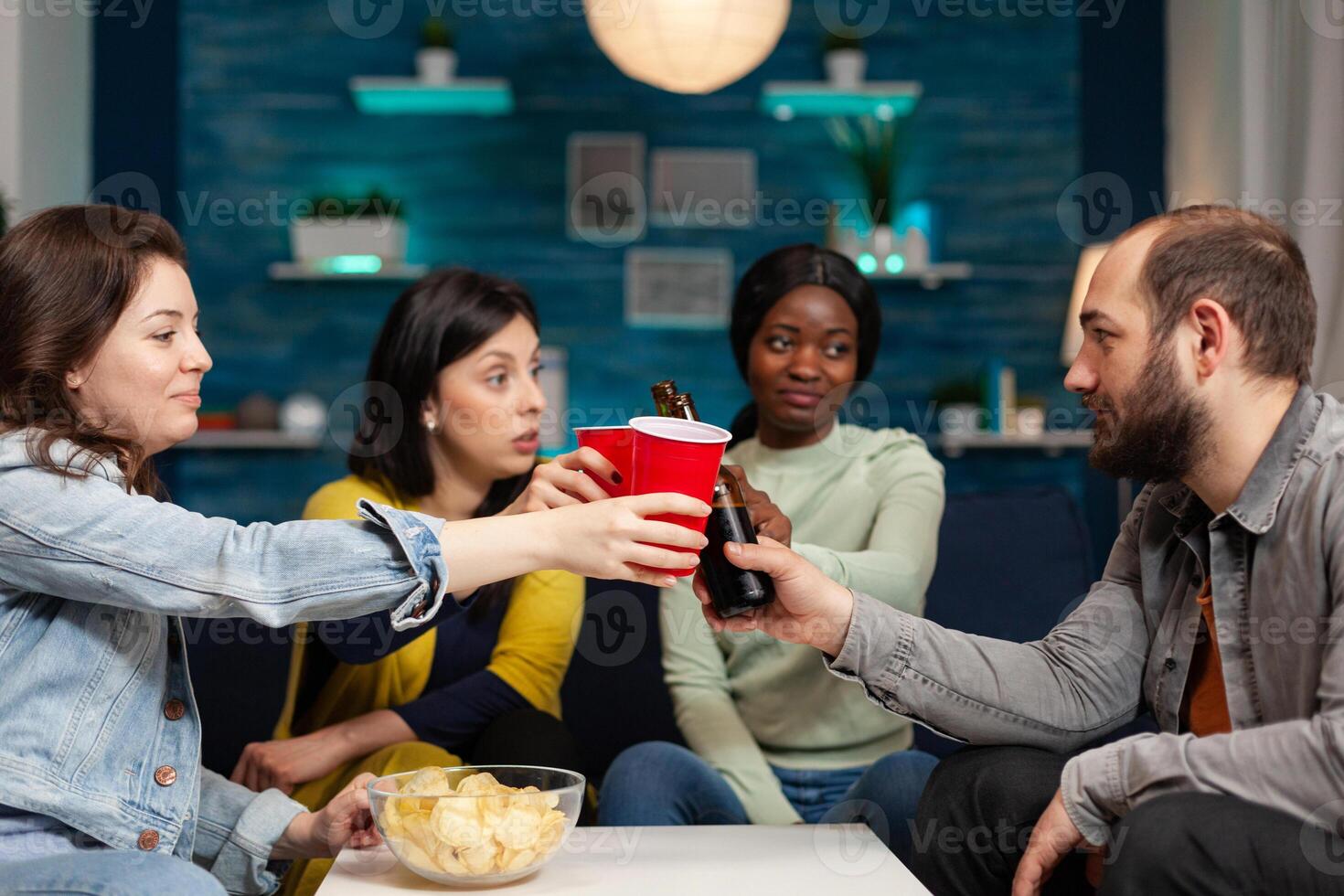 Happy cheerful multi ethnic friends clinking beer while having fun and entertaining themselfGroup of multiracial people spending time together sitting on couch late at night in living room. photo