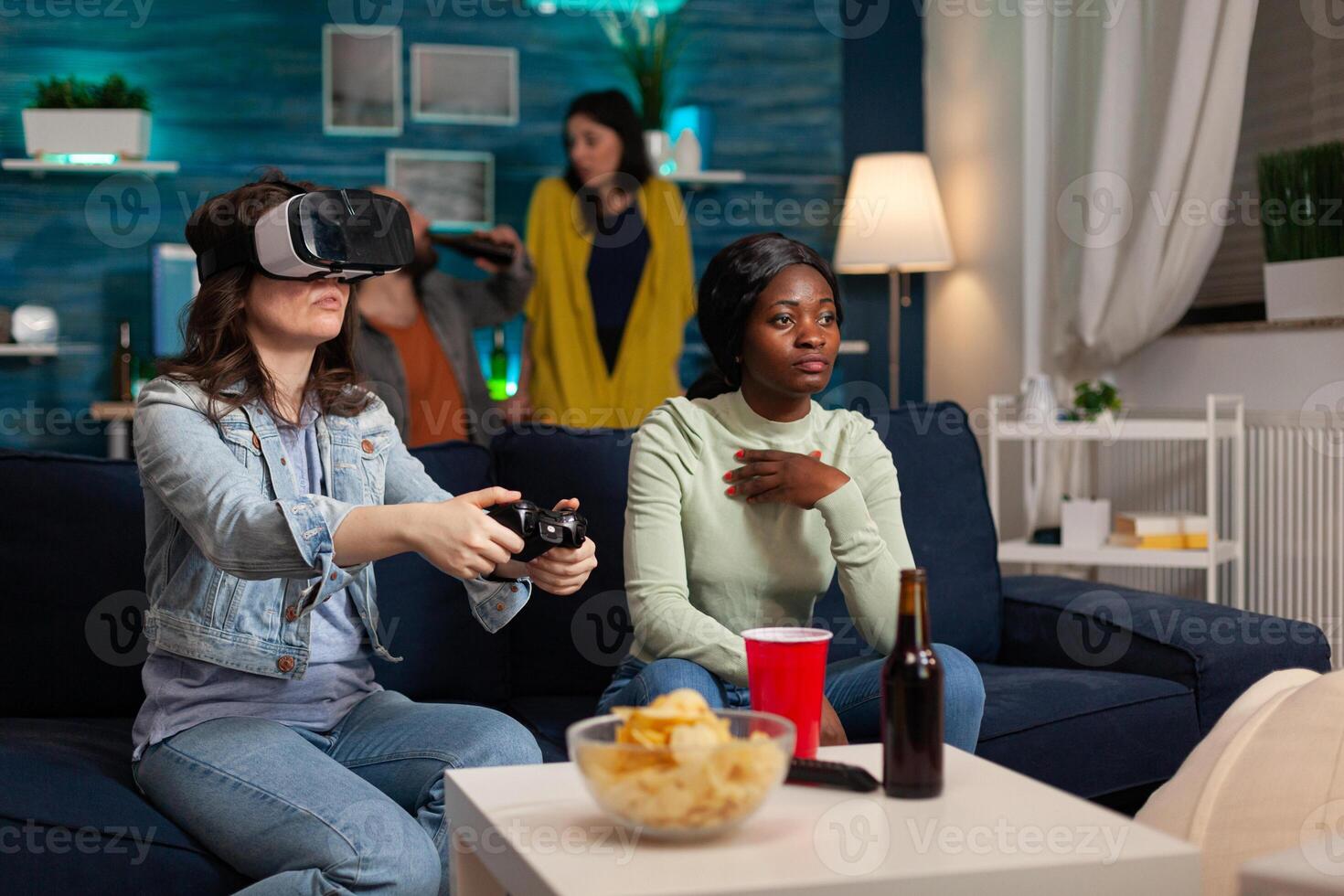 Mixed race friends having online virtual games competition wearing vr headset, using wireless controller, hanging out late in the evening sitting on sofa drinking beer and enjoying snacks. photo