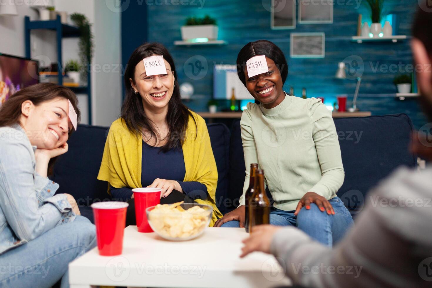 Mixed race friends playing Guess who game with sticky papers attached on forehead. Group of multiracial people having fun, laughing together while sitting on sofa in living room late at night. photo
