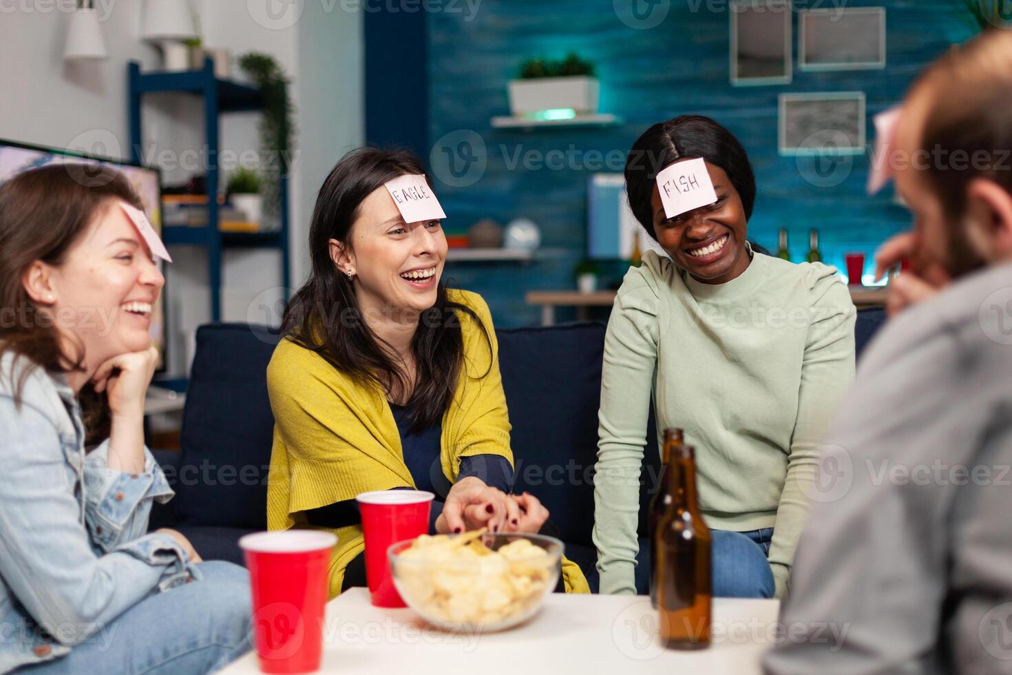 Mixed race friends gathering in living room playing guess who game using sticky notes. Mixed race people having fun, laughing together while sitting on sofa late at night during funny party. photo