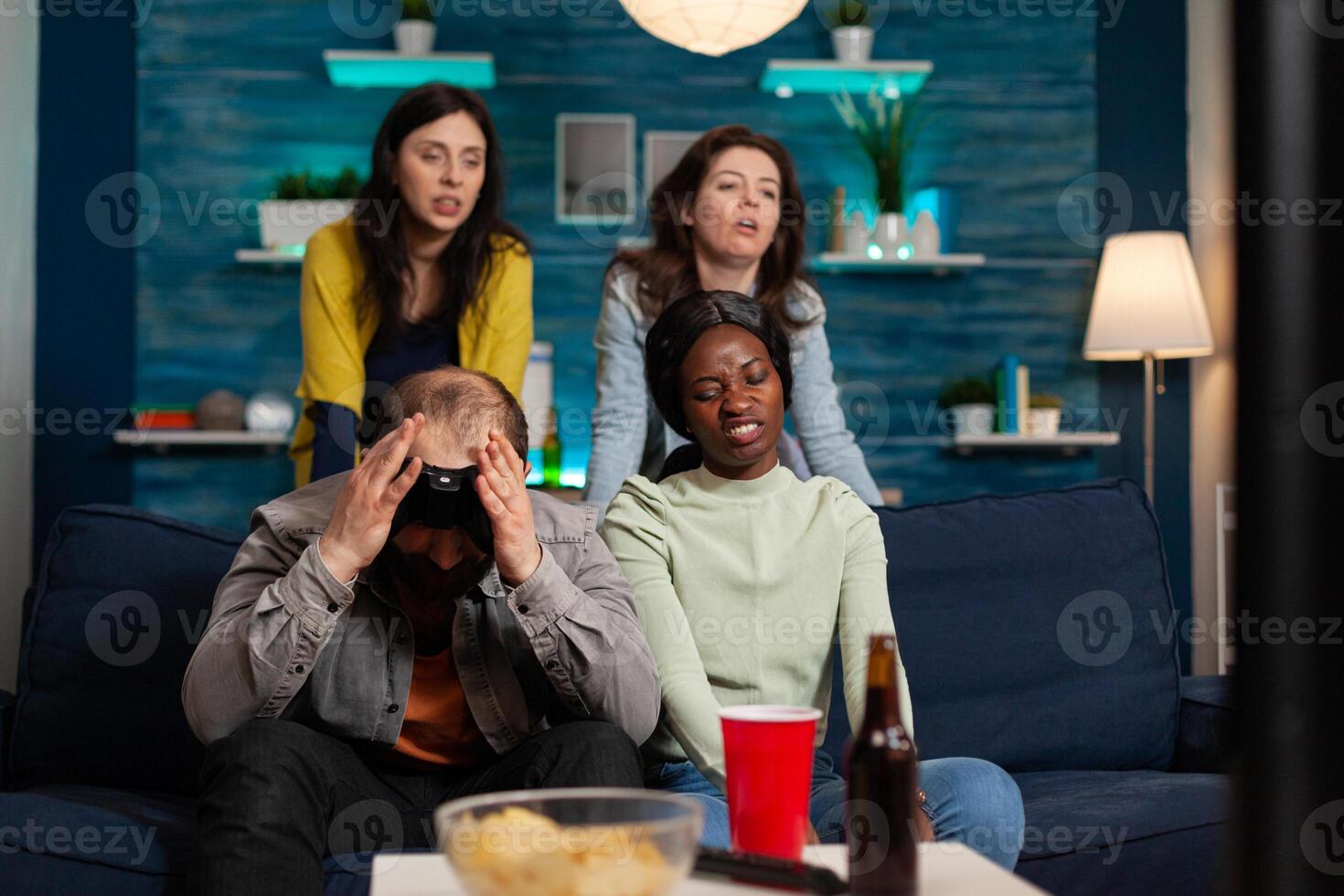 Angry man and multi ethnic friends upset after losing game competition, bonding and sitting on couch after drinking beer. Mixed race group of people hanging out together having fun late at night in living room photo
