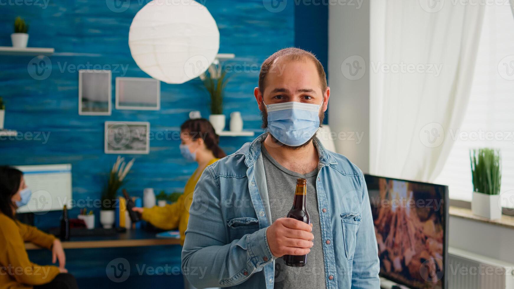 Man with protection mask looking at camera holding bottle of beer ready for party with friends in living room respecting social distance in global pandemic. People socializing during covid 19 outbreak photo