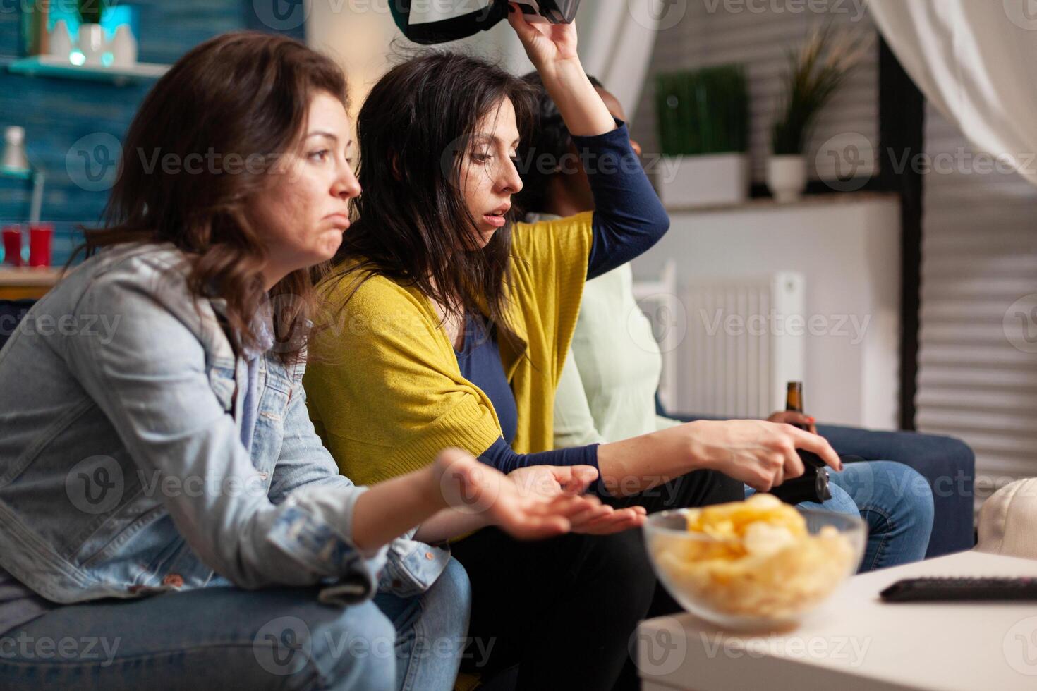 Multi ethnic friends upset after losing virtual reality gaming competition sitting on couch. Mixed race group of people hanging out together having fun late at night in living room photo