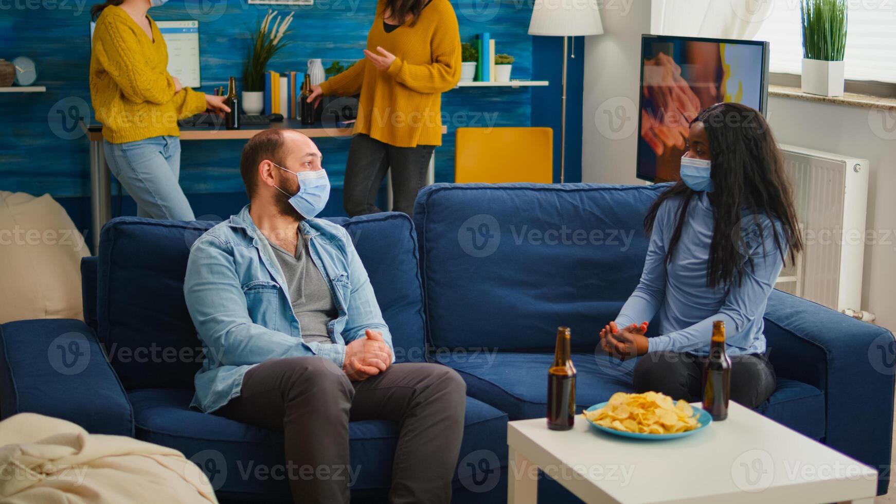 Diverse friends discussing during new normal party wearing protection mask, socializing keeping distance sitting on sofa drinking beer and eating snaks. Group of multi ethnic people enjoying free time photo