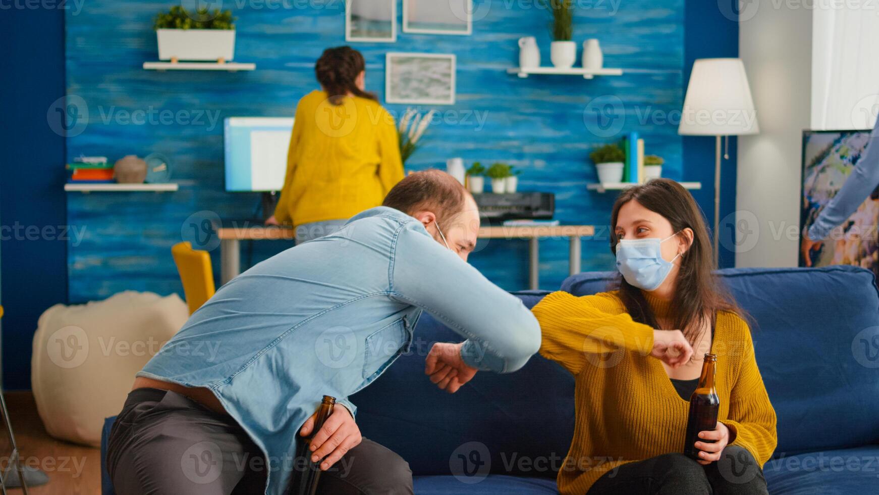 Friends with protection mask socializing during party sitting on sofa touching elbow keeping social distancing greeting each other preventing spread of coronavirus, holding beer bottles in living room photo