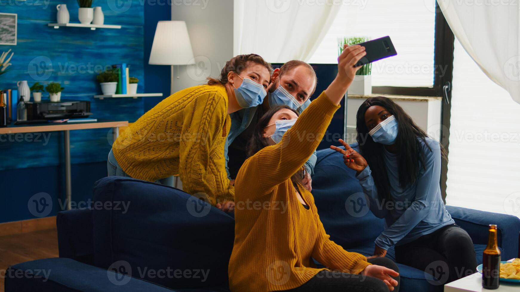 Multiracial friends taking selfie with face masks during covid 19 outbreak, new normal lifestyle concept with people having fun in living room respecting social distance to prevent virus spread photo