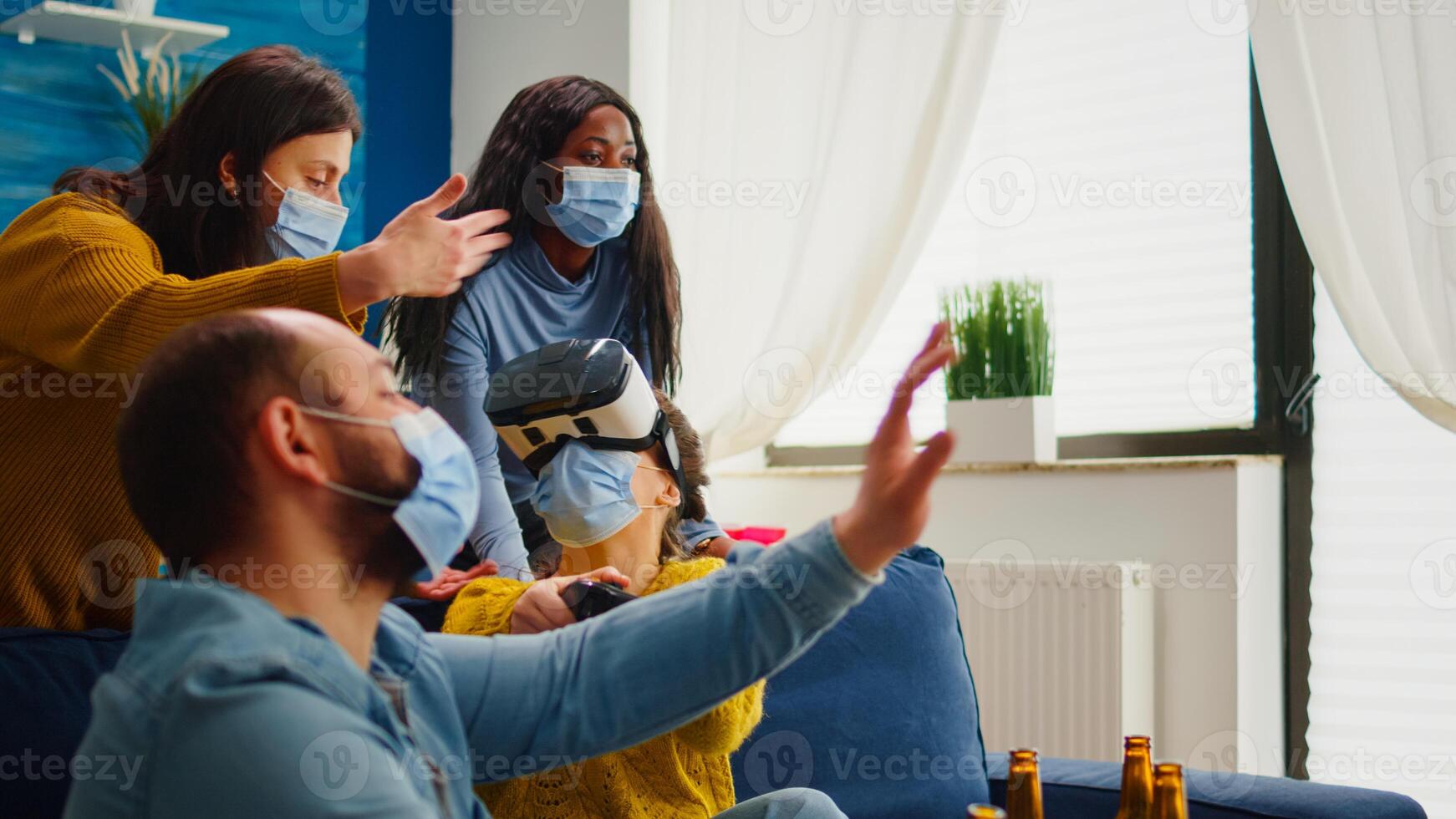 Friends supporting woman during game playing with virtual reality goggles wearing protection mask agaist covid19 virus. Diverse people having fun, laughing, drinking beer at new normal party photo