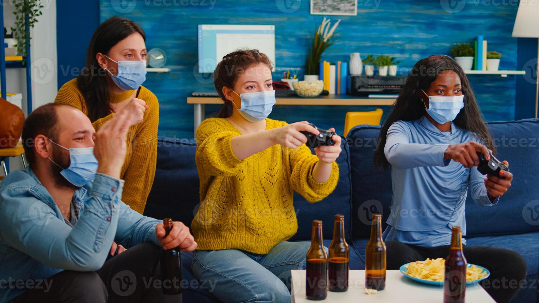 Mixed race women with protection mask playing game holding wireless controller enjoying new normal party relaxing with friends. Diverse people keeping social distancing against global pandemic photo