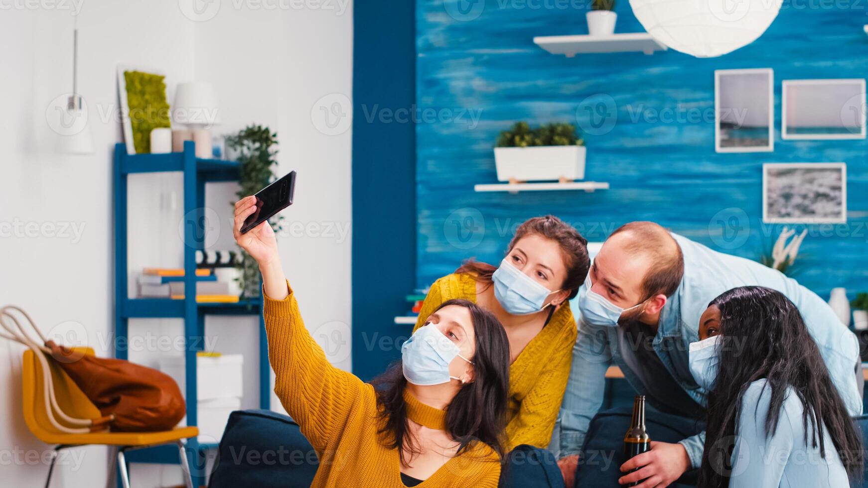 Multi-ethnnic friends with protective masks partying with beer taking selfie during covid 19 outbreak, new normal lifestyle concept with people having fun in living room respecting social distance photo