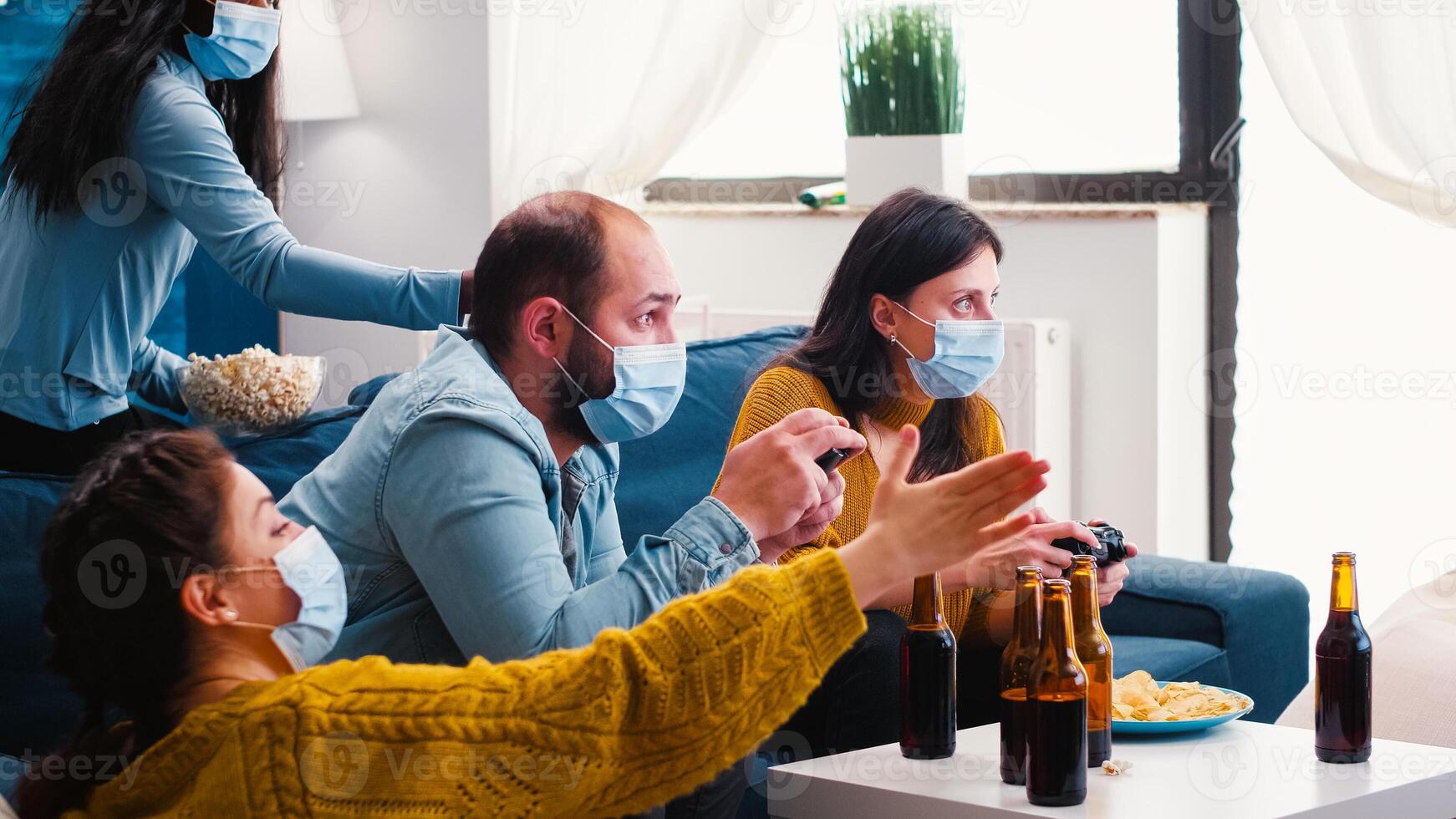 Multiracial people celebrate winning game in home living room with joystick wearing face mask keeping social distancing in time of corona outbreak. Diverse friends enoying beer and chips. photo